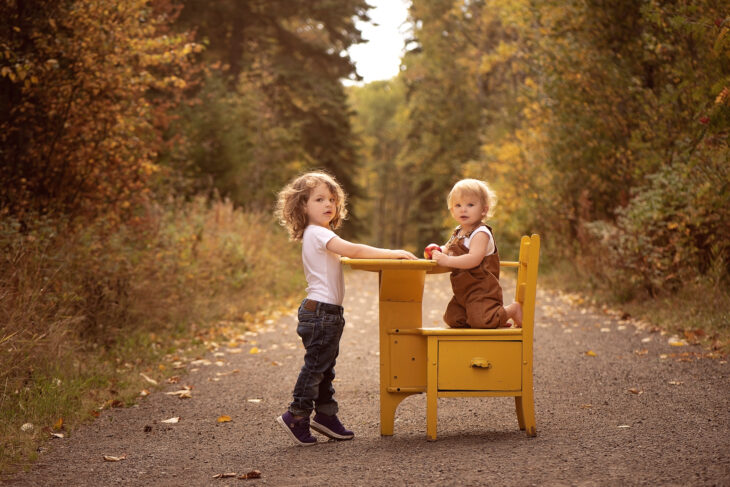 in a forest one 1 year old sits at a yellow desk with an apple and their sibling standing on their tiptoes in purple shoes